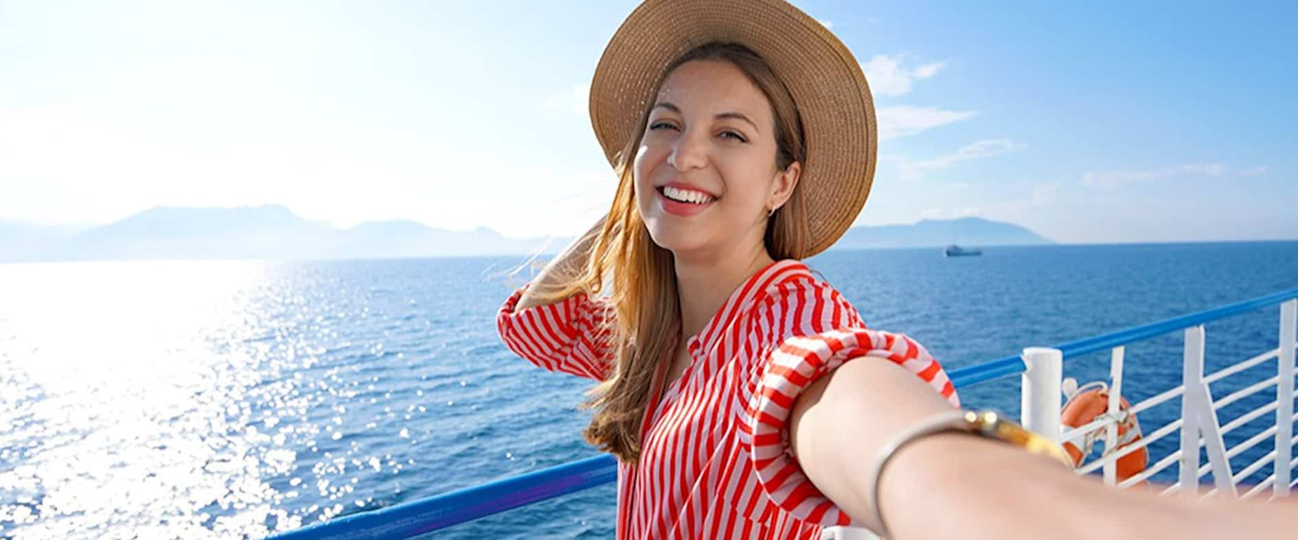 Glücklich lachende junge Frau mit Sonnenhut an Deck eines Schiffs, im Hintergrund das glitzernde Meer und strahlender Sonnenschein.