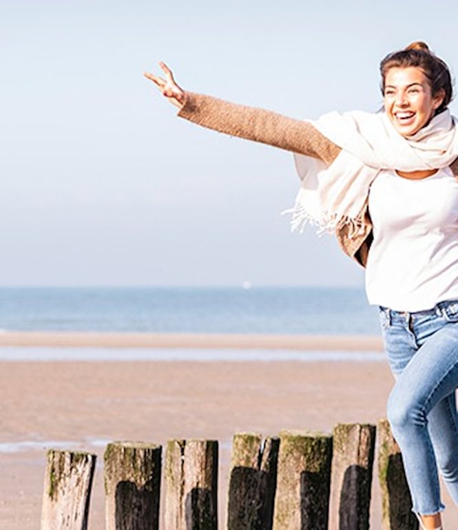 Junge Frau lacht glücklich am Meer, mit Sonne im Gesicht und Wind im Haar - ein Moment voller Lebensfreude.