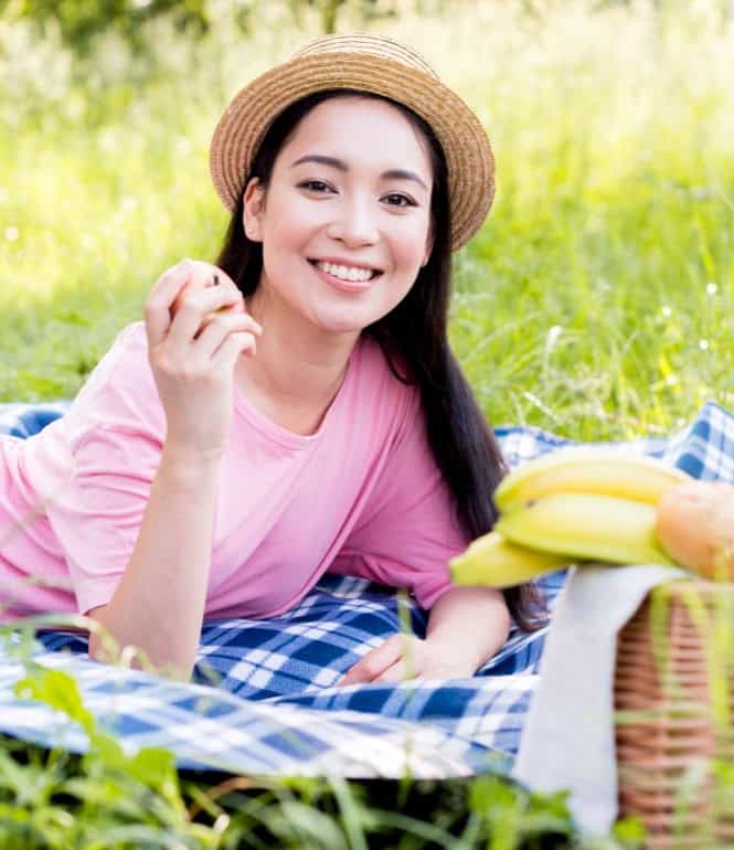 Junge Frau liegt lächelnd auf einer Picknickdecke mit Picknickkorb – entspannter Frühlingsmoment unter freiem Himmel.