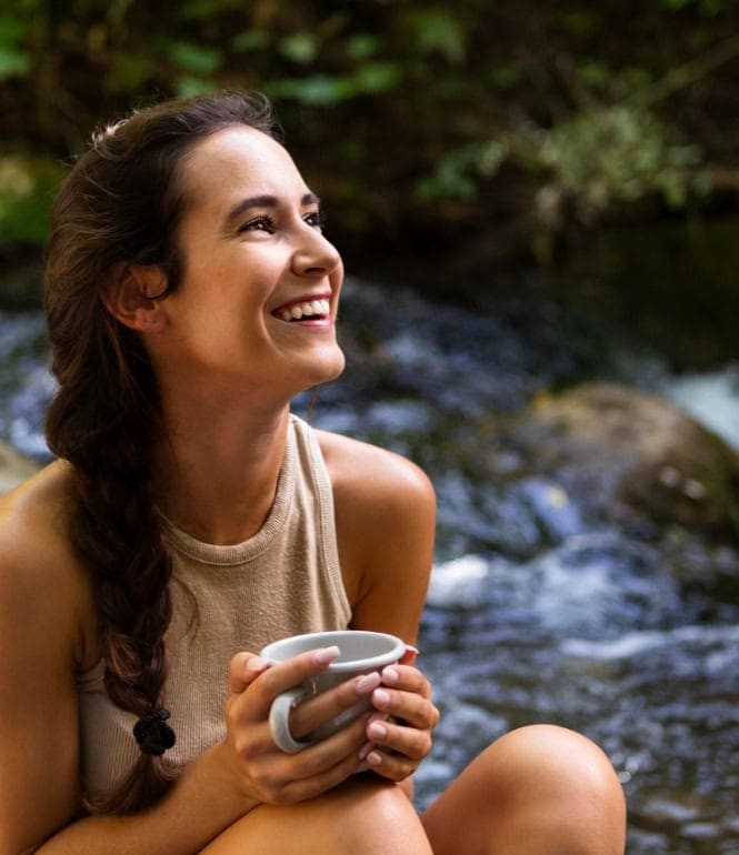 Junge Frau sitzt lächelnd an einem Fluss – sie wirkt glücklich und erfüllt.