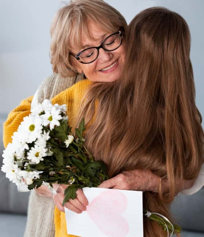 Tochter umarmt ihre Mutter liebevoll, Mutter lächelt und hält einen Blumenstrauß und eine Karte in den Händen.