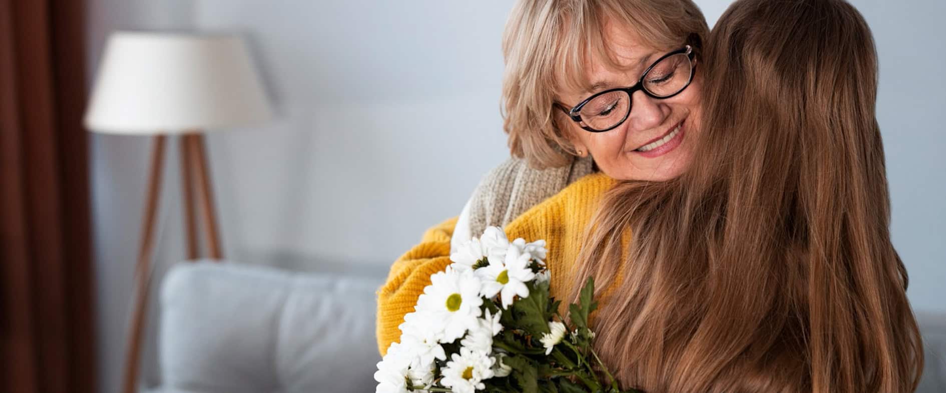 Tochter umarmt ihre Mutter liebevoll, Mutter lächelt und hält einen Blumenstrauß und eine Karte in den Händen.