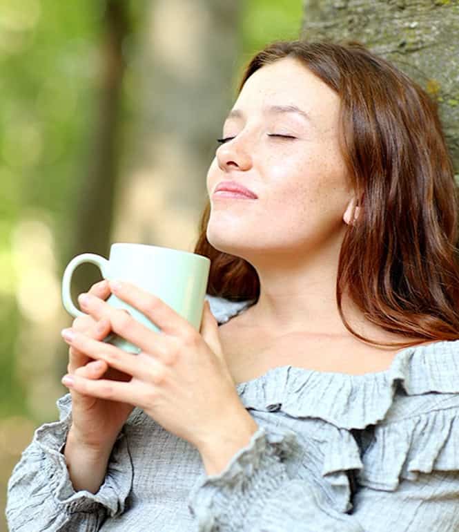 Junge Frau lehnt entspannt an einem Baum, trinkt genussvoll Kaffee oder Tee mit geschlossenen Augen