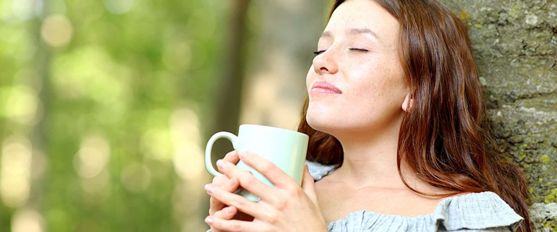 Junge Frau lehnt entspannt an einem Baum, trinkt genussvoll Kaffee oder Tee mit geschlossenen Augen