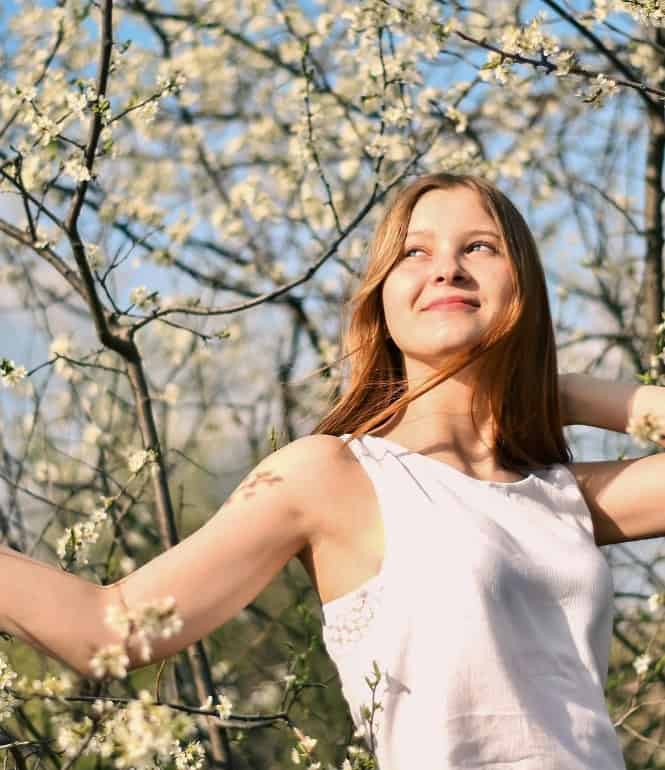 Lächelnde junge Frau im weißen Kleid vor blühenden Bäumen und blauem Himmel, Symbol für Frühlingsgefühle, Neuanfang und Lebensfreude