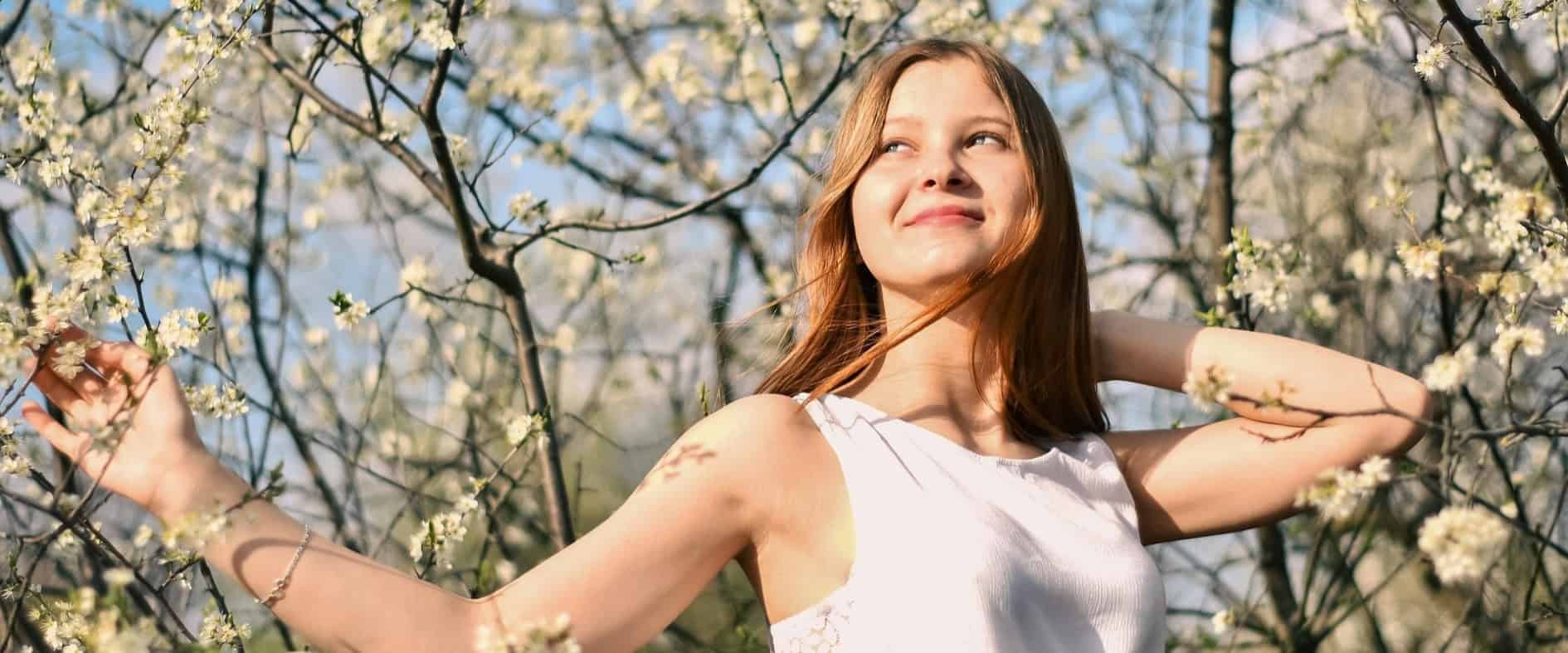 Lächelnde junge Frau im weißen Kleid vor blühenden Bäumen und blauem Himmel, Symbol für Frühlingsgefühle, Neuanfang und Lebensfreude
