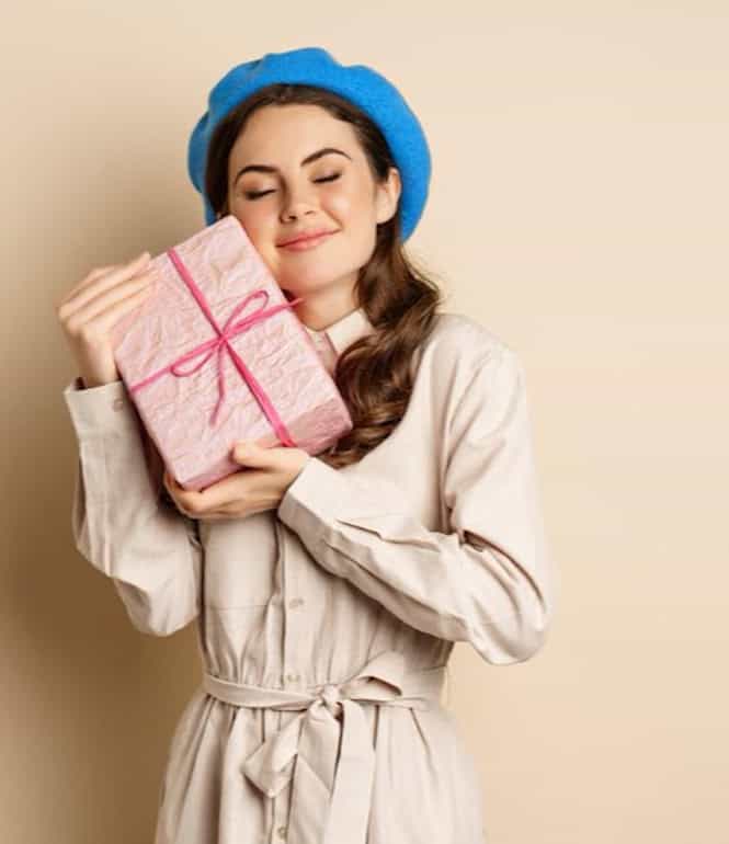 Junge Frau mit blauer Baskenmütze hält ein eingepacktes Geschenk nah an sich und lächelt mit geschlossenen Augen.