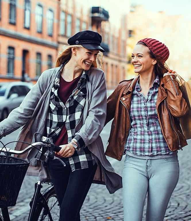 Zwei junge Frauen lachen beim Stadtbummel, eine schiebt ein Fahrrad, die andere trägt Einkaufstüten – Szene in einer lebendigen, fahrradfreundlichen Innenstadt.