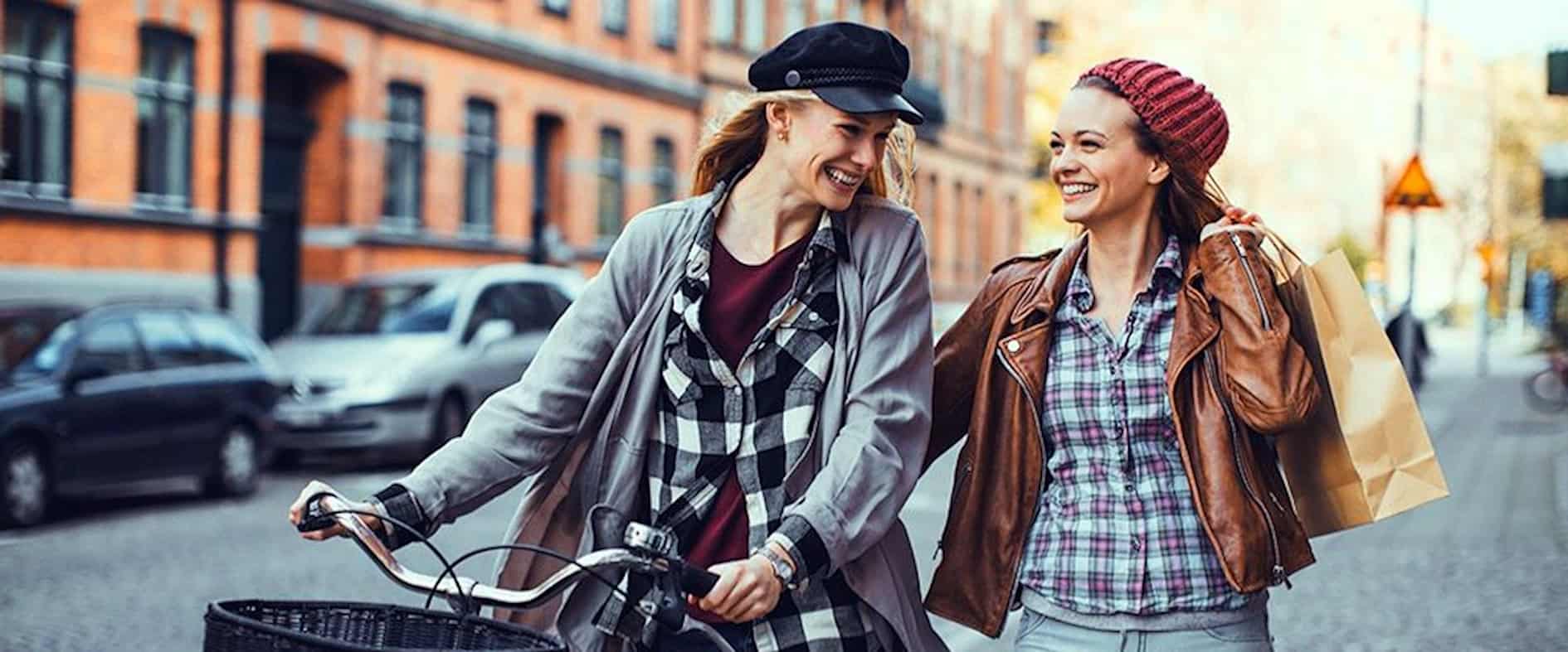 Zwei junge Frauen lachen beim Stadtbummel, eine schiebt ein Fahrrad, die andere trägt Einkaufstüten – Szene in einer lebendigen, fahrradfreundlichen Innenstadt.