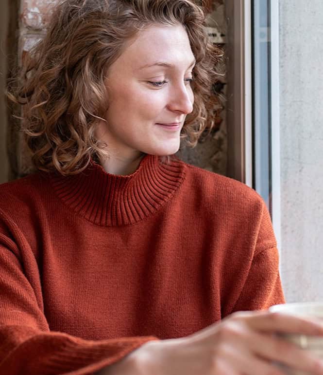 Junge Frau in rotem Pullover sitzt mit einer Tasse am Fenster und lächelt nachdenklich – Symbolbild für berufliche Selbstreflexion und die Suche nach Erfüllung.