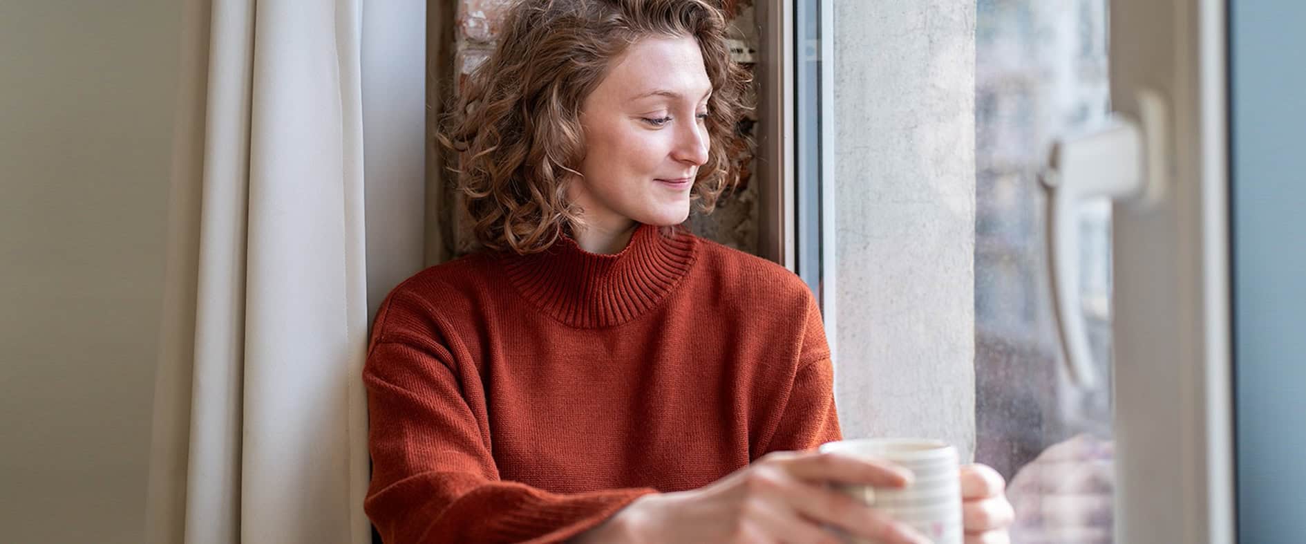 Junge Frau in rotem Pullover sitzt mit einer Tasse am Fenster und lächelt nachdenklich – Symbolbild für berufliche Selbstreflexion und die Suche nach Erfüllung.