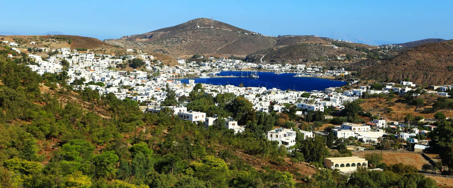 Blick über den Hafen von Patmos mit strahlend blauem Himmel, ruhigem Meer und sanften Hügeln im Hintergrund
