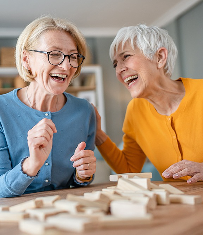 Zwei ältere Frauen lachen gemeinsam beim Spielen am Tisch, Symbolbild für Lebensfreude, soziale Verbundenheit und aktives Altern.