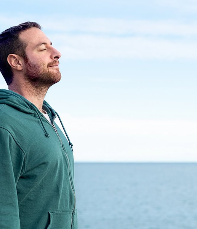Ein Mann steht entspannt am Meer, schließt die Augen und atmet tief ein – Symbol für innere Ruhe und bewusste Achtsamkeit.
