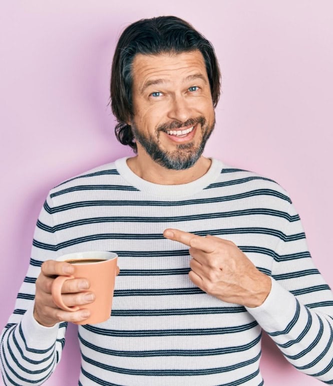 Mann mit gestreiftem Pullover und Kaffeetasse vor rosa Hintergrund, lächelt und zeigt auf seine Tasse.
