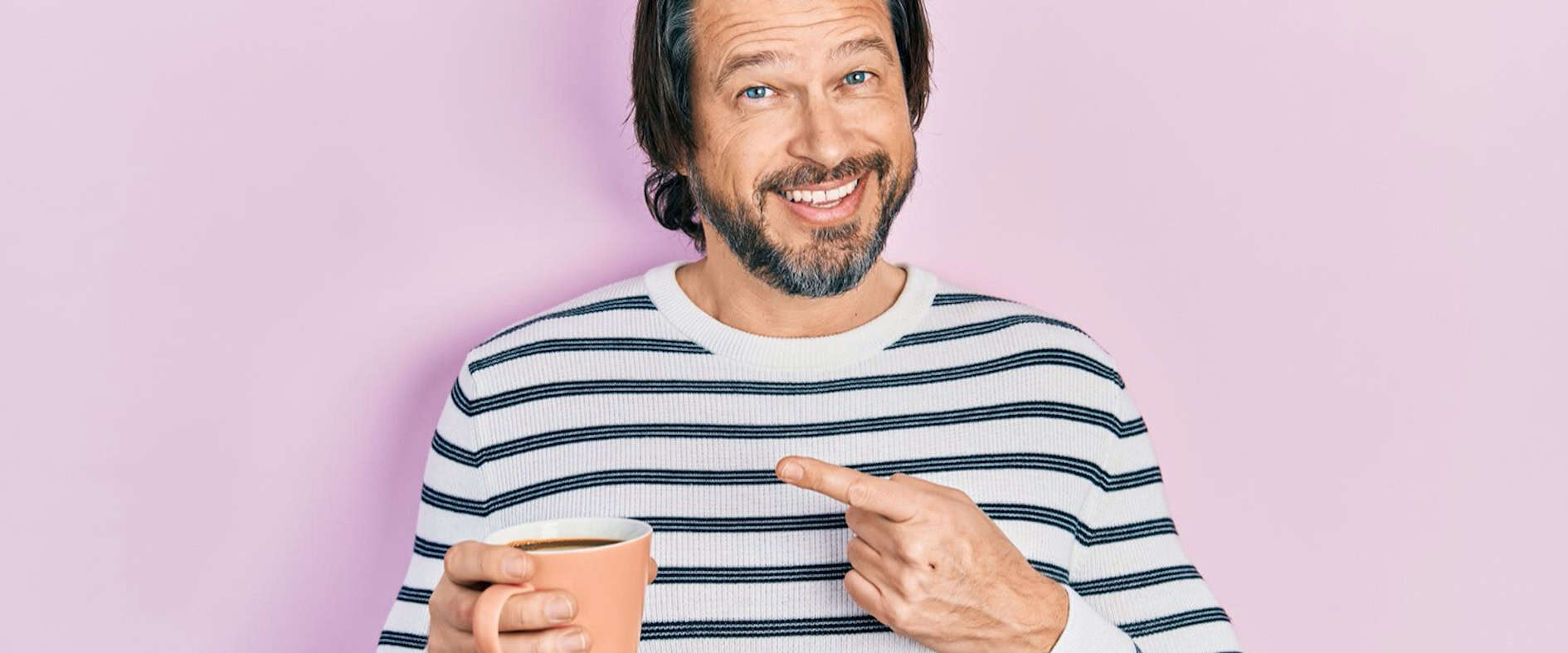 Mann mit gestreiftem Pullover und Kaffeetasse vor rosa Hintergrund, lächelt und zeigt auf seine Tasse.
