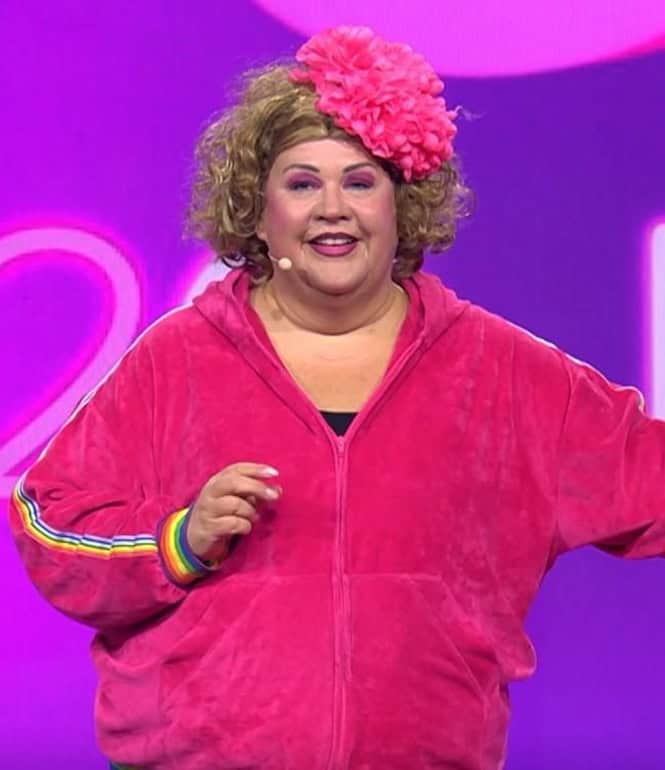 Cindy aus Marzahn auf der Bühne in pinkem Jogginganzug mit großer Blume im Haar, Mikrofon in der Hand, vor pink-violettem Bühnenhintergrund.