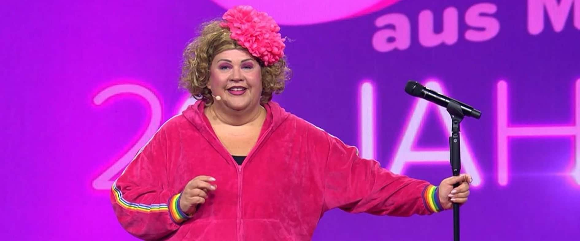 Cindy aus Marzahn auf der Bühne in pinkem Jogginganzug mit großer Blume im Haar, Mikrofon in der Hand, vor pink-violettem Bühnenhintergrund.