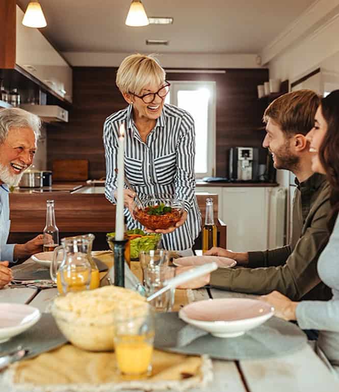 Vier Erwachsene und eine ältere Frau lachen gemeinsam beim Essen an einem Esstisch in einer gemütlichen Küche – Symbol für Glück und Zusammenhalt.