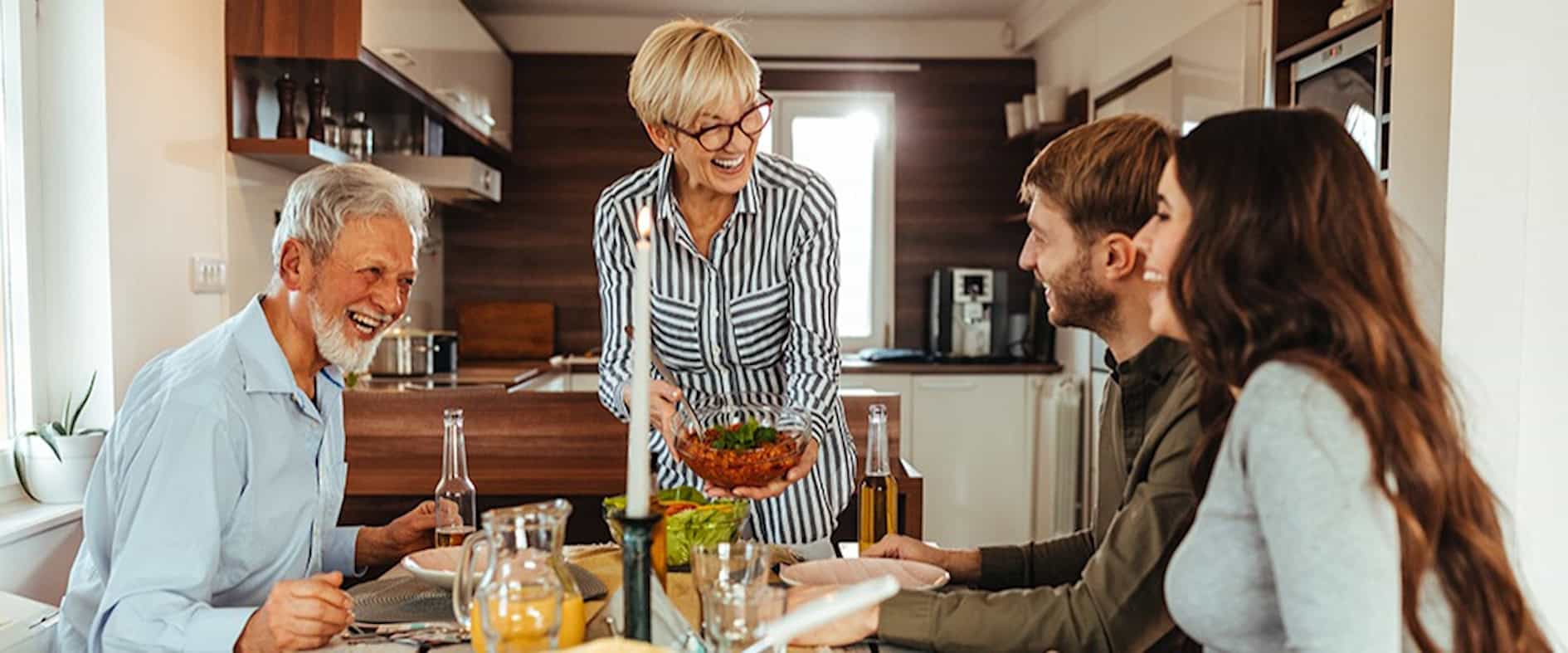 Vier Erwachsene und eine ältere Frau lachen gemeinsam beim Essen an einem Esstisch in einer gemütlichen Küche – Symbol für Glück und Zusammenhalt.