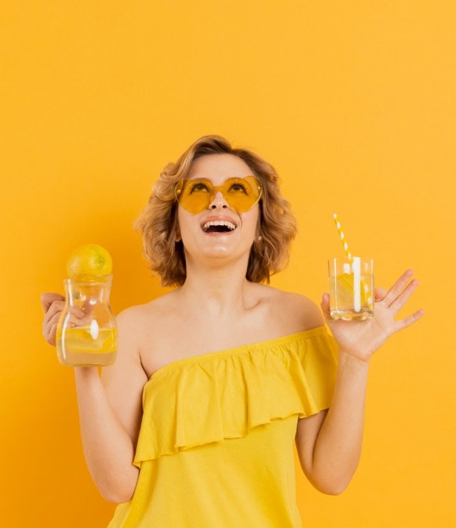 Lächelnde Frau mit gelbem Outfit und Sonnenbrille hält ein Glas und eine Karaffe mit Zitronenwasser vor orangefarbenem Hintergrund.