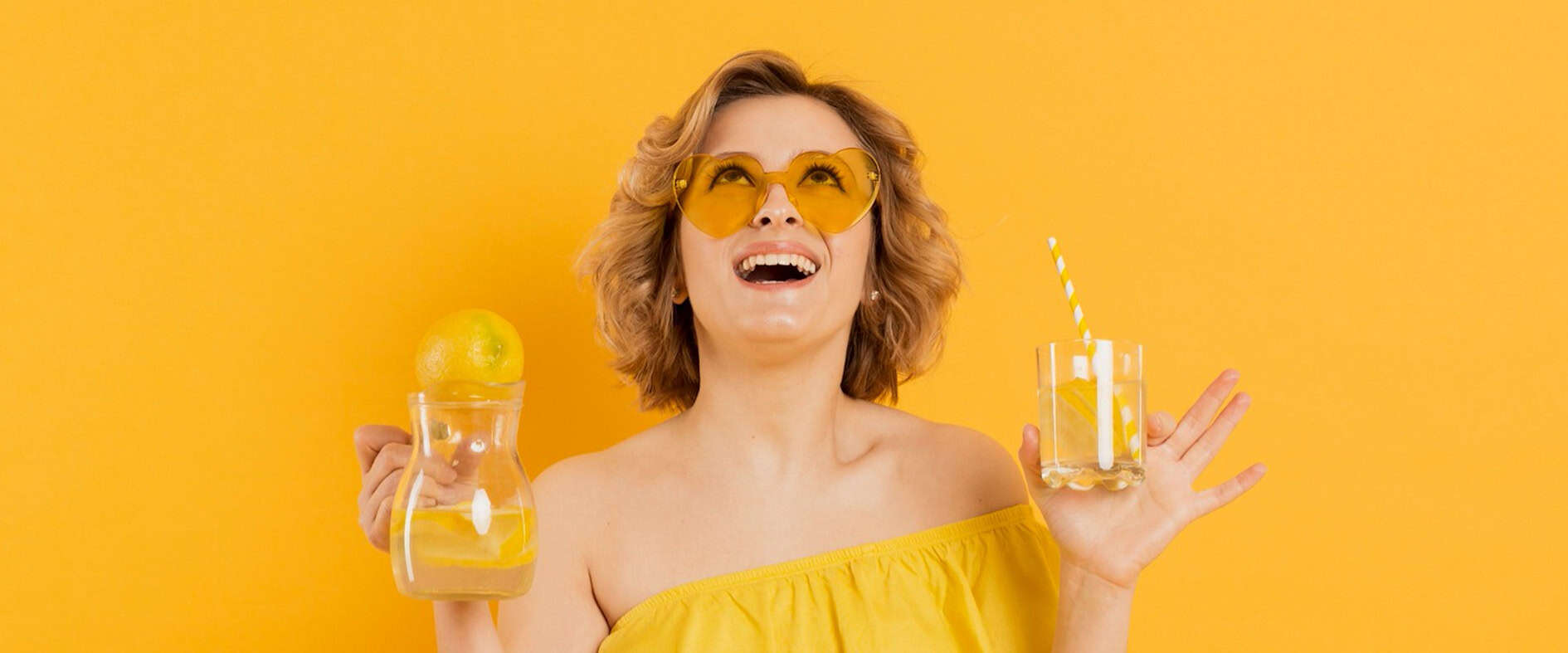 Erfrischend durch den Sommer: Die besten Durstlöscher Lächelnde Frau mit gelbem Outfit und Sonnenbrille hält ein Glas und eine Karaffe mit Zitronenwasser vor orangefarbenem Hintergrund.