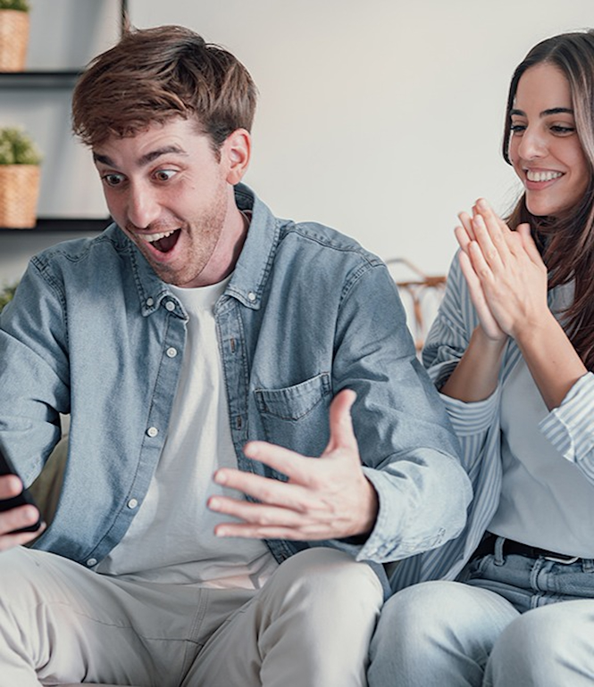 Mann und Frau freuen sich gemeinsam auf dem Sofa über eine Nachricht auf dem Smartphone, symbolisch für einen doppelten Lottogewinn.