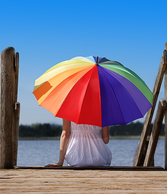 Person sitzt auf einem Holzsteg am Wasser unter einem großen, bunten Sonnenschirm.