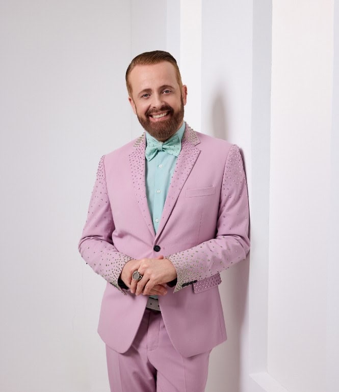 Porträt von Ross Antony im rosa Anzug mit türkisfarbener Fliege, lächelnd vor hellem Hintergrund – Promofoto zum Interview und Album „100 Jahre gute Laune“.