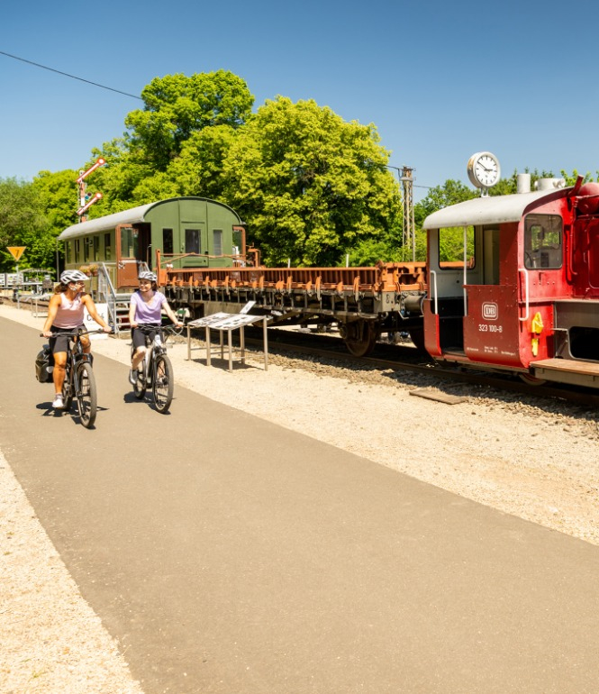 Zwei Radfahrerinnen fahren bei sonnigem Wetter auf einem asphaltierten Radweg entlang einer historischen roten Deutz-Kleinlok und alten Güterwaggons in Pronsfeld, Eifel.