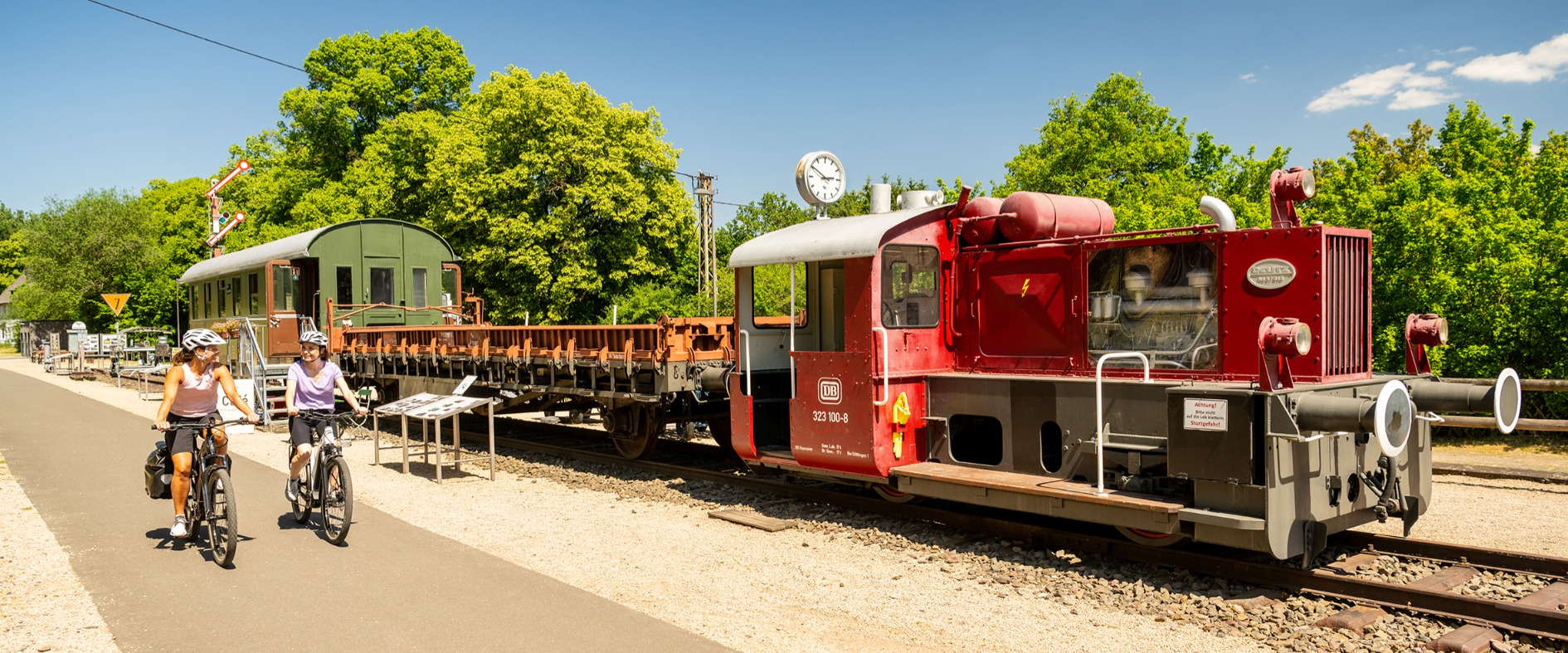 Zwei Radfahrerinnen fahren bei sonnigem Wetter auf einem asphaltierten Radweg entlang einer historischen roten Deutz-Kleinlok und alten Güterwaggons in Pronsfeld, Eifel.