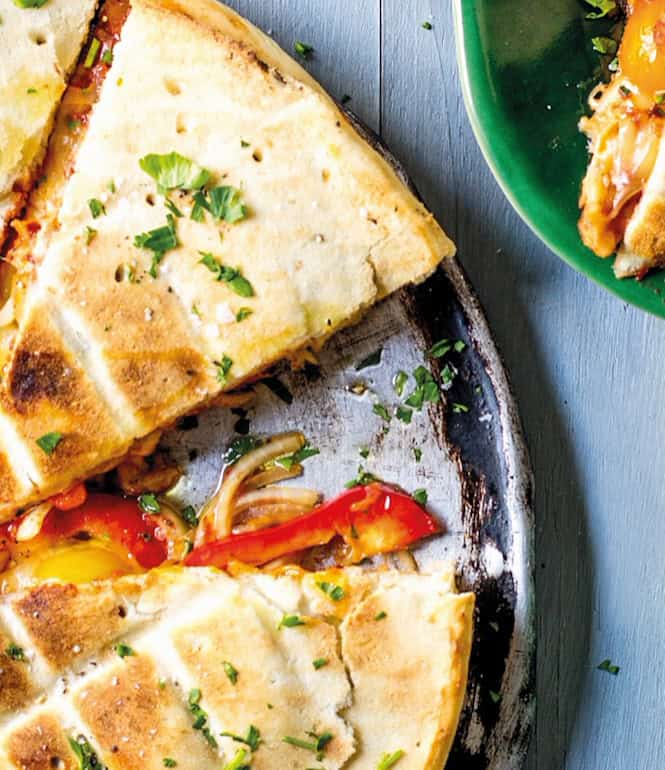 Gefüllte Pizza vom Grill, in Stücke geschnitten, serviert auf einem Blech mit frischen Kräutern bestreut, daneben ein Teller mit einem Pizzastück.