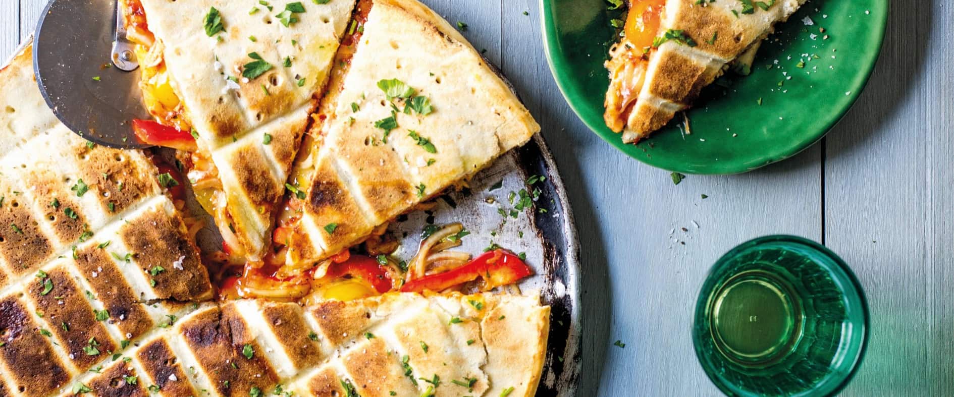 Gefüllte Pizza vom Grill, in Stücke geschnitten, serviert auf einem Blech mit frischen Kräutern bestreut, daneben ein Teller mit einem Pizzastück.