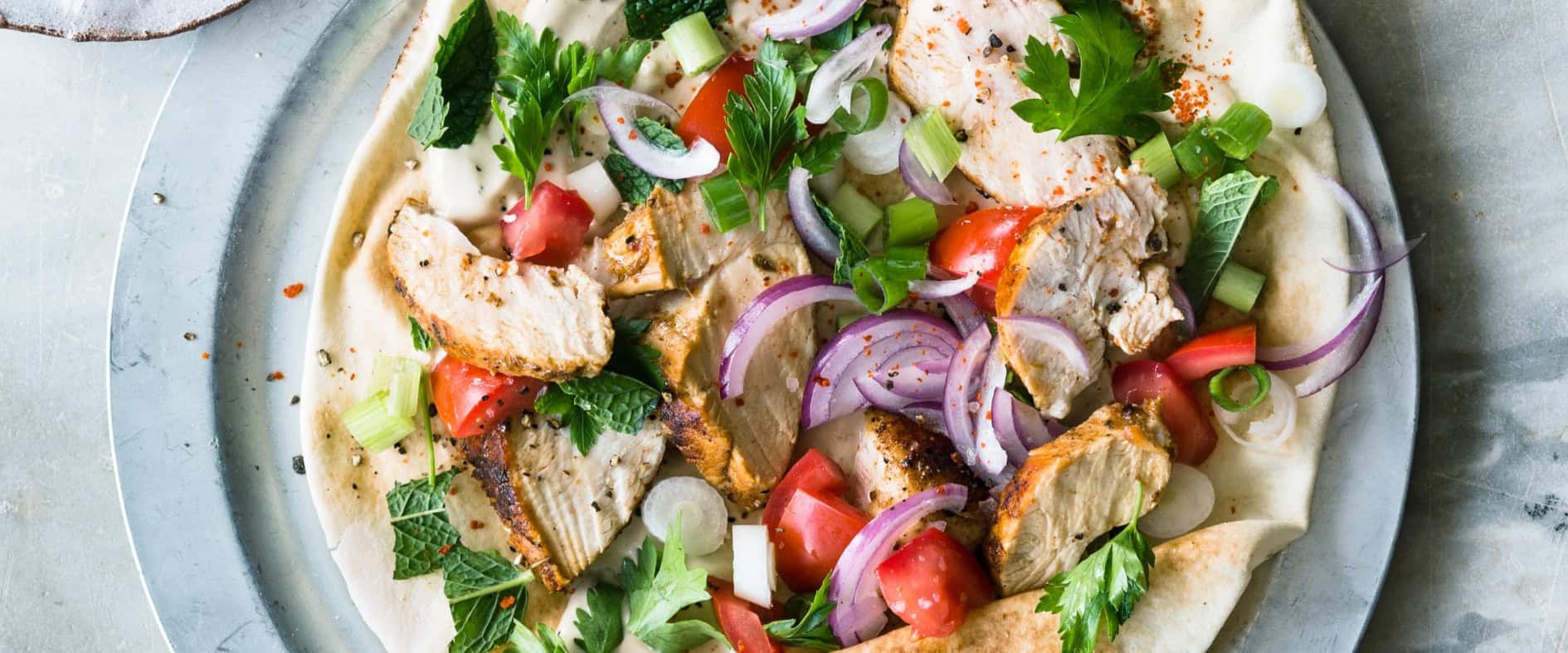 Fladenbrot mit gegrilltem Shawarma-Hähnchen, Tomaten, roten Zwiebeln, Frühlingszwiebeln, frischen Kräutern und Tahina-Dip, angerichtet auf einem Teller.