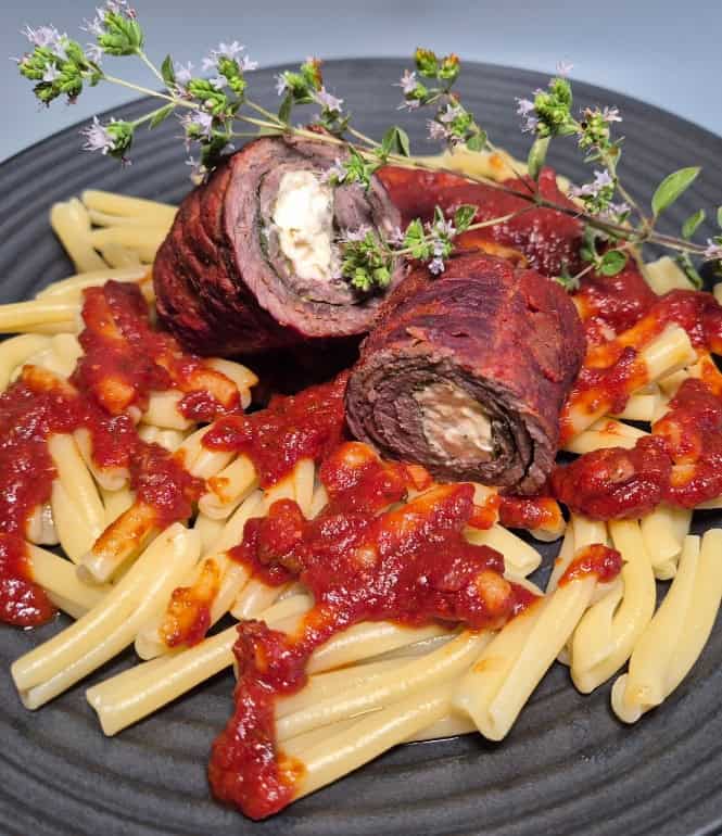 Gefüllte mediterrane Rinderroulade mit Schafskäse und Spinat, serviert auf Pasta mit würziger Tomatensauce und frischen Kräutern garniert.