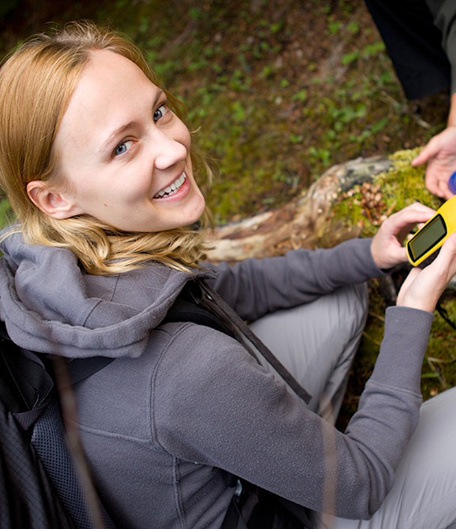 Junge Frau beim Geocaching im Wald, hält ein GPS-Gerät in der Hand und lächelt in die Kamera, daneben wird ein gefundener Cache geöffnet.