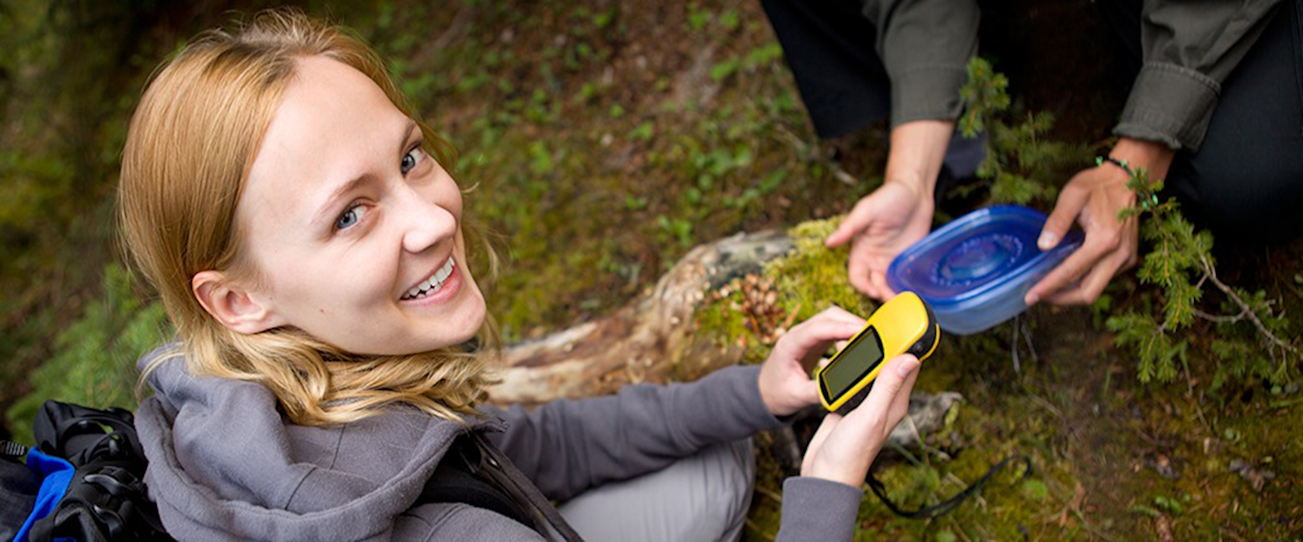 Junge Frau beim Geocaching im Wald, hält ein GPS-Gerät in der Hand und lächelt in die Kamera, daneben wird ein gefundener Cache geöffnet.