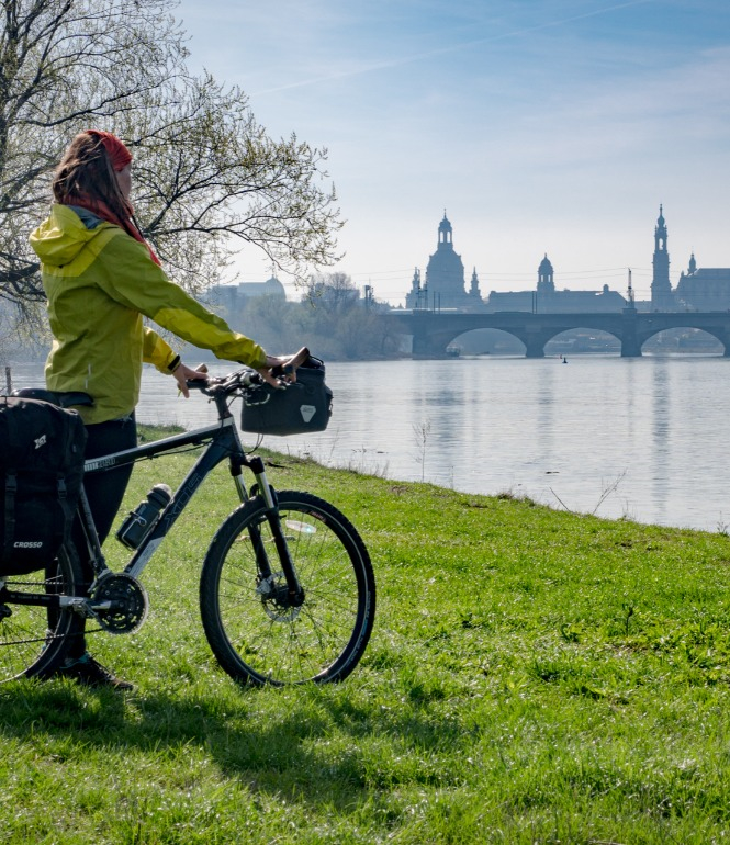 Radfahrerin mit Gepäck steht am Elbufer und blickt auf die Dresdner Altstadt mit Augustusbrücke im Hintergrund.