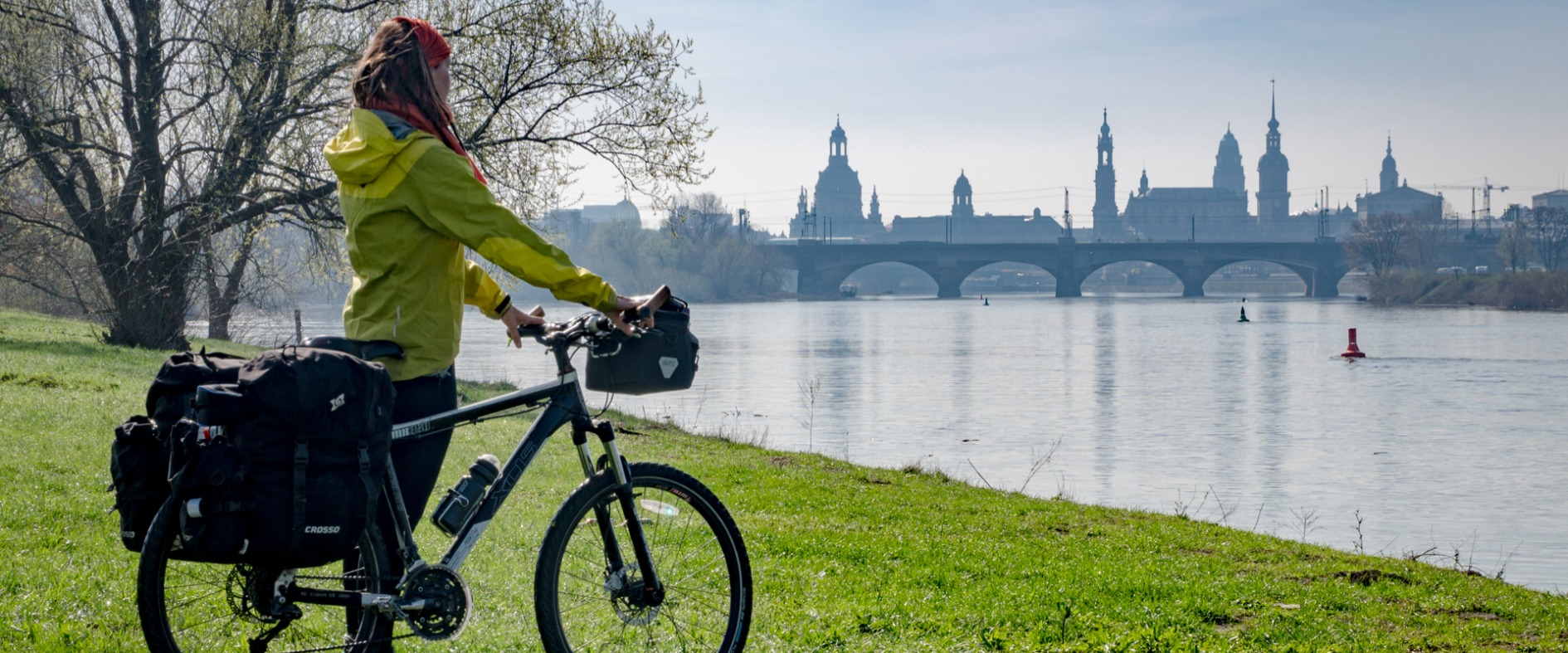 Radfahrerin mit Gepäck steht am Elbufer und blickt auf die Dresdner Altstadt mit Augustusbrücke im Hintergrund.