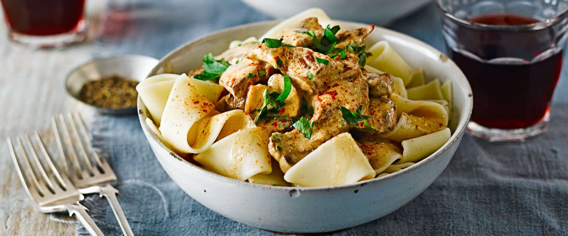 Schüsseln mit cremigem Rinder-Stroganoff und Pappardelle, garniert mit frischer Petersilie, serviert auf einem gedeckten Tisch.
