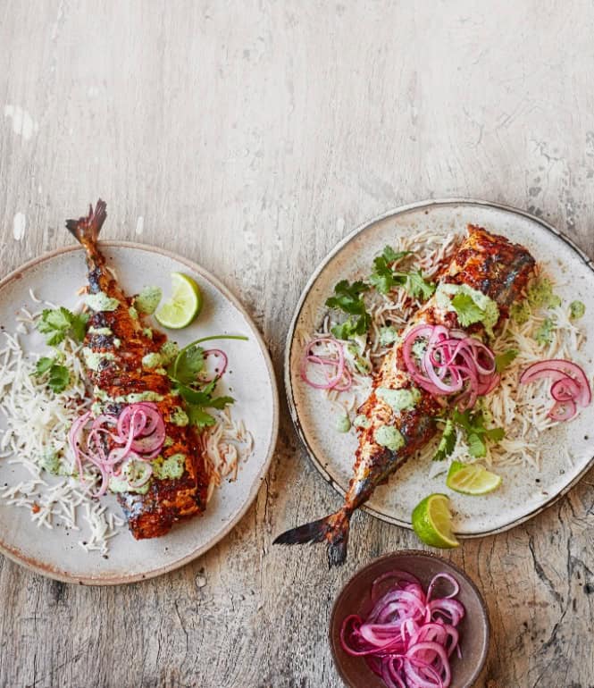 Gegrillte Masala-Makrelen auf Basmatireis, garniert mit eingelegten roten Zwiebeln, Koriander, Limettenspalten und grüner Joghurt-Sauce, serviert auf zwei Tellern.