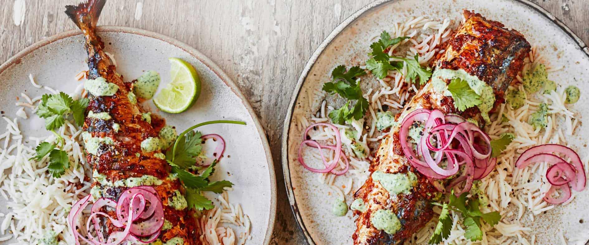 Gegrillte Masala-Makrelen auf Basmatireis, garniert mit eingelegten roten Zwiebeln, Koriander, Limettenspalten und grüner Joghurt-Sauce, serviert auf zwei Tellern.