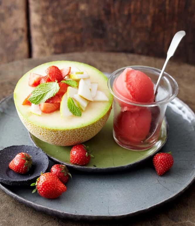 Halbe Galiamelone gefüllt mit Erdbeeren, Birne und Minze, serviert mit einem Glas Wassermelonensorbet und frischen Erdbeeren auf einem Teller.