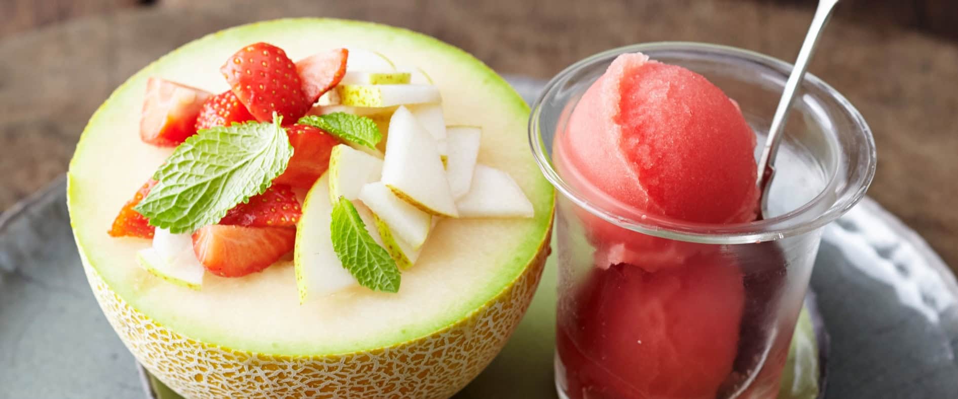 Halbe Galiamelone gefüllt mit Erdbeeren, Birne und Minze, serviert mit einem Glas Wassermelonensorbet und frischen Erdbeeren auf einem Teller.