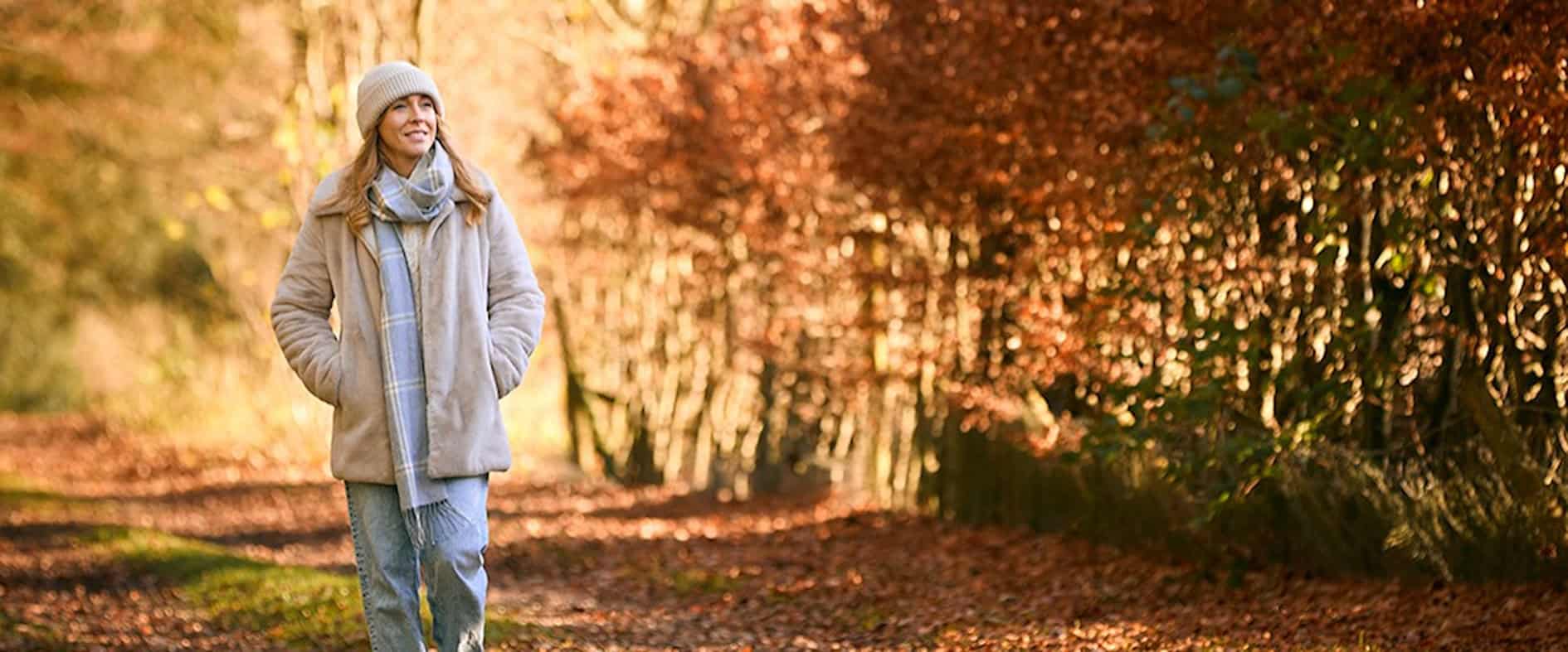 Frau geht entspannt in warmer Kleidung durch einen herbstlichen Waldweg.