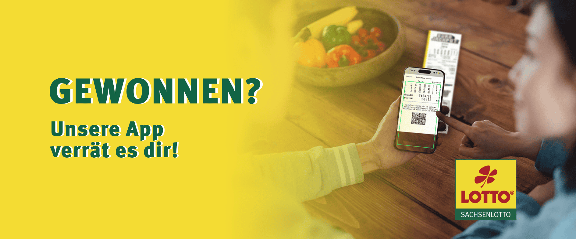 Zwei Personen sitzen an einem Tisch und scannen mit dem Smartphone einen Lottoschein. Auf dem Bildschirm ist der QR-Code der Spielquittung zu sehen. Darüber steht der Text: „Gewonnen? Unsere App verrät es dir!“ – Werbung für die Sachsenlotto-App mit Scan-Funktion.