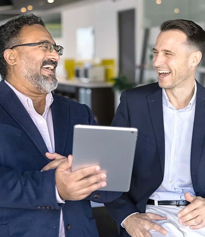 Zwei Männer in Businesskleidung lachen gemeinsam bei einem Gespräch und halten ein Tablet.