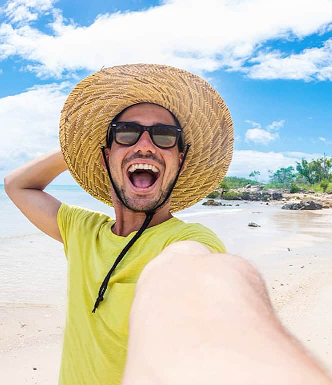 Junger Mann mit Sonnenhut und Sonnenbrille macht ein Selfie am Strand.