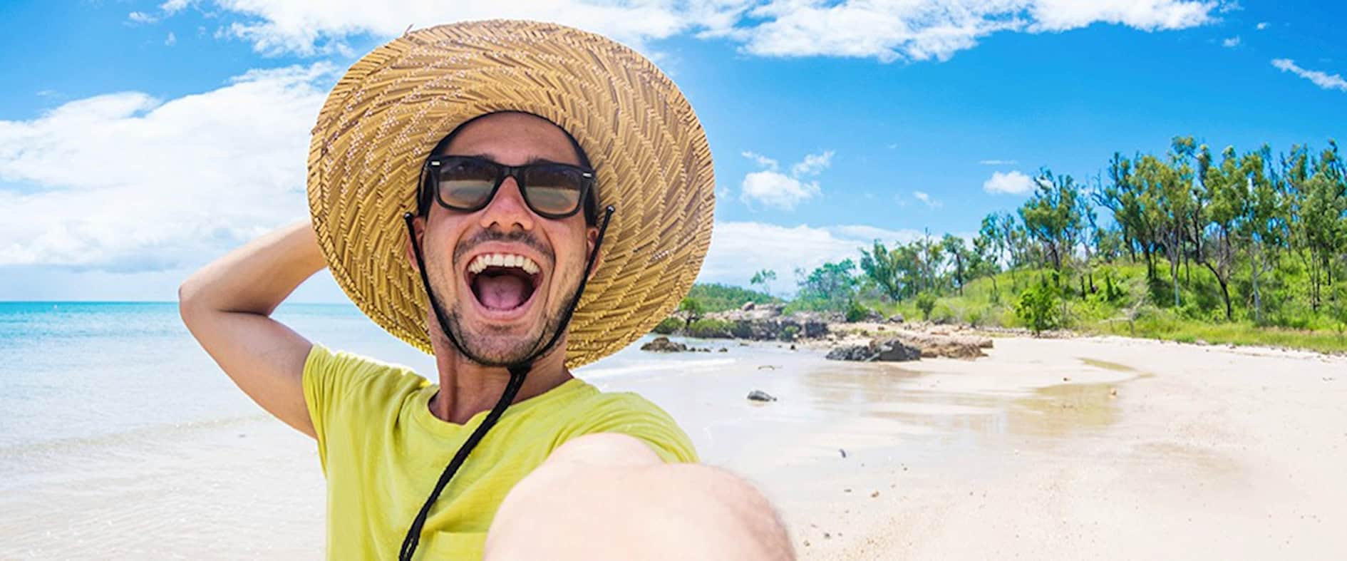 Junger Mann mit Sonnenhut und Sonnenbrille macht ein Selfie am Strand.
