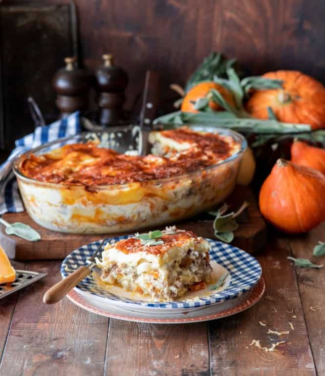 Kürbis-Lasagne mit Hackfleisch, cremiger Sauce und geschmolzenem Käse, serviert auf einem Teller, im Hintergrund eine Auflaufform mit frischer Lasagne und Herbstdeko mit Kürbissen.